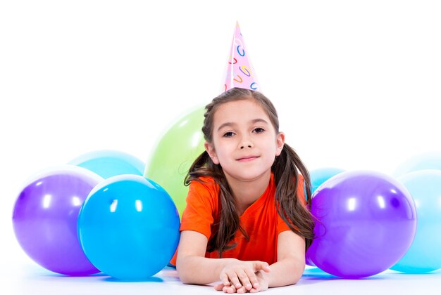 Feliz niña sonriente en camiseta naranja tirada en el suelo con globos de colores - aislado en un blanco