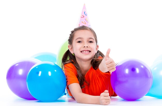 Feliz niña sonriente en camiseta naranja tirada en el suelo con globos de colores y mostrando los pulgares hacia arriba - aislado en blanco