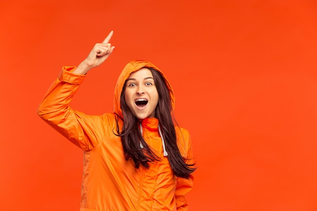 La feliz niña sonriente posando en el estudio en otoño chaqueta naranja apuntando hacia arriba aislado en rojo. Emociones humanas positivas. Concepto del clima frío. Conceptos de moda femenina