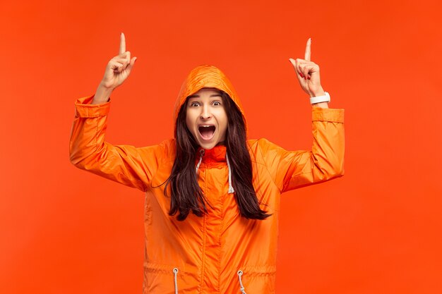 La feliz niña sonriente posando en el estudio en otoño chaqueta naranja apuntando hacia arriba aislado en rojo. Emociones humanas positivas. Concepto del clima frío