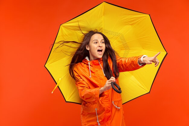 La feliz niña sonriente posando en el estudio en otoño chaqueta naranja y apuntando a la izquierda aislada en rojo.