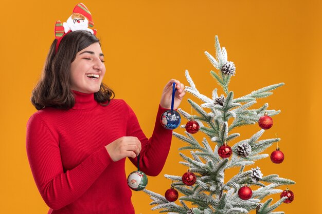 Feliz niña en suéter de navidad con diadema divertida junto a un árbol de navidad sobre fondo naranja