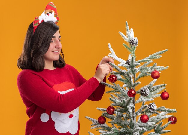 Feliz niña en suéter de Navidad con diadema divertida de pie junto a un árbol de Navidad colgando bolas de Navidad sobre la pared naranja