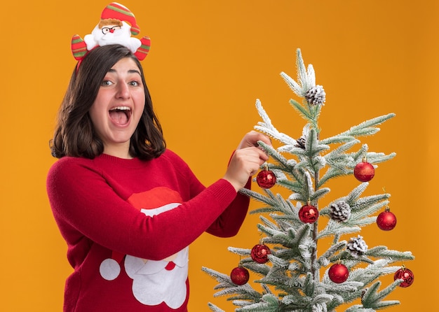 Foto gratuita feliz niña en suéter de navidad con diadema divertida con una sonrisa en la cara de pie junto a un árbol de navidad colgando bolas de navidad sobre la pared naranja