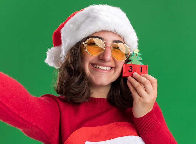 Feliz niña en suéter de Navidad con gorro de Papá Noel y gafas sosteniendo cubos de juguete con fecha de año nuevo con gran sonrisa en la cara de pie sobre la pared verde