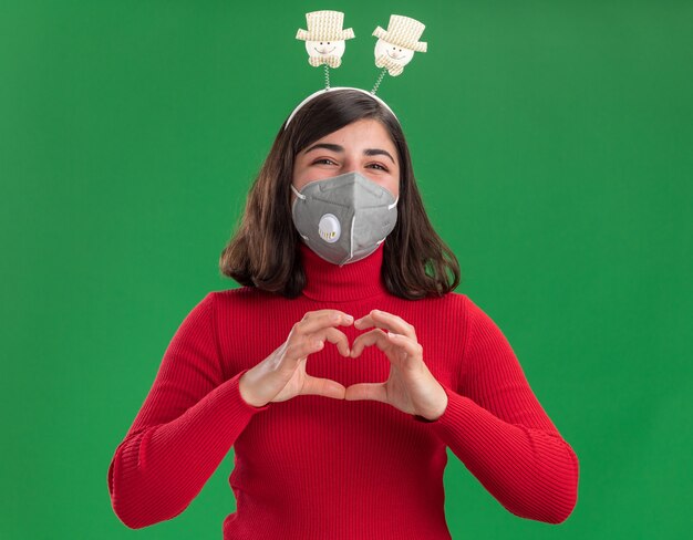 Feliz niña en suéter rojo con diadema divertida con máscara protectora facial haciendo gesto de corazón con los dedos de pie sobre la pared verde