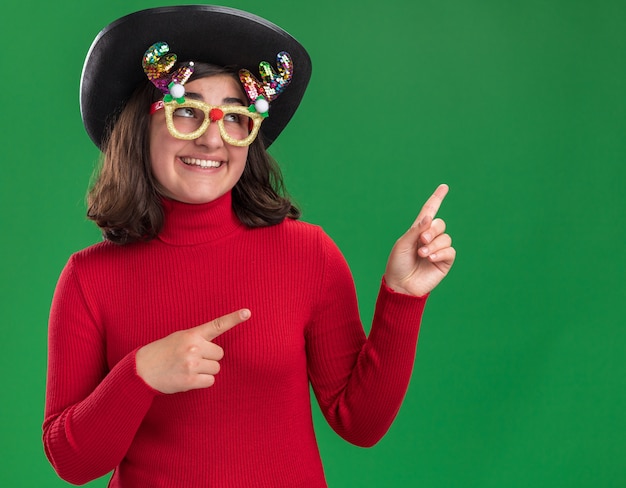 Feliz niña de suéter rojo con gafas divertidas y sombrero negro mirando hacia arriba