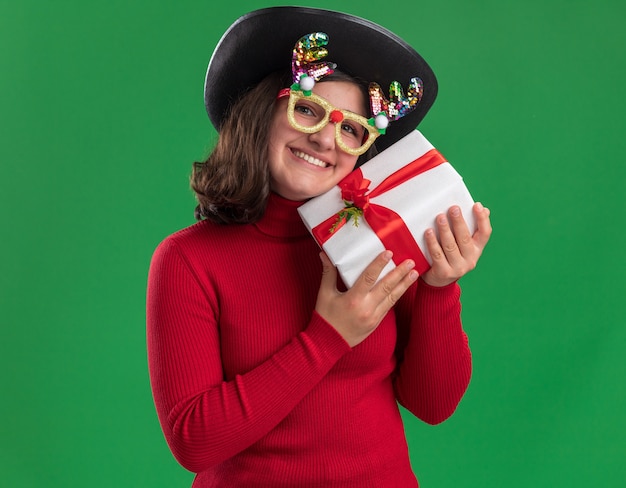 Feliz niña de suéter rojo con gafas divertidas y sombrero negro sosteniendo un presente mirando a la cámara sonriendo alegremente de pie sobre fondo verde