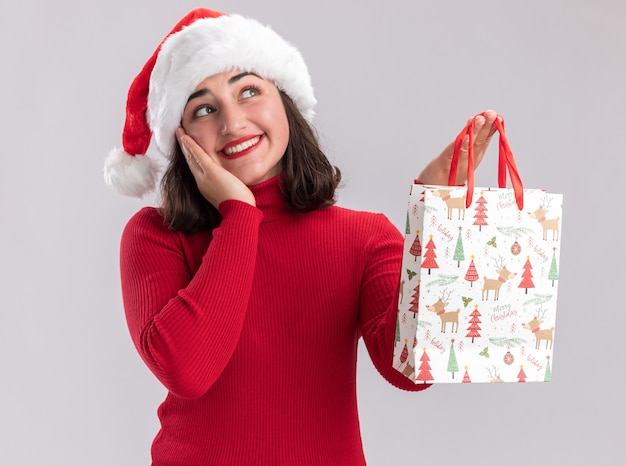 Feliz niña de suéter rojo y gorro de Papá Noel con bolsa de papel de colores con regalos de Navidad mirando hacia arriba con una sonrisa en la cara de pie sobre fondo blanco.