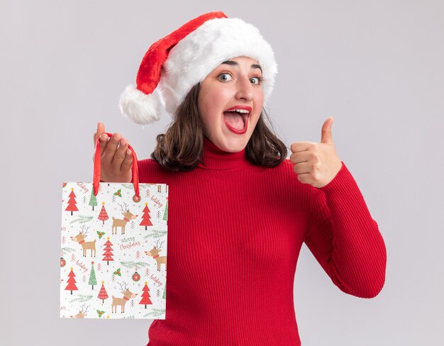 Feliz niña en suéter rojo y gorro de Papá Noel con bolsa de papel de colores con regalos de Navidad mirando a la cámara con una sonrisa en la cara mostrando los pulgares para arriba sobre fondo blanco.