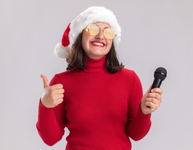 Feliz niña de suéter rojo y gorro de Papá Noel con gafas sosteniendo el micrófono mirando a la cámara sonriendo alegremente mostrando los pulgares para arriba sobre fondo blanco.