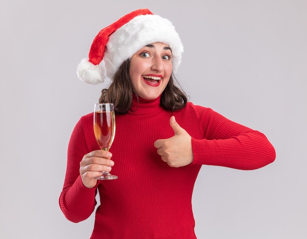 Feliz niña de suéter rojo y gorro de Papá Noel sosteniendo una copa de champán mirando a la cámara sonriendo alegremente mostrando los pulgares para arriba sobre fondo blanco