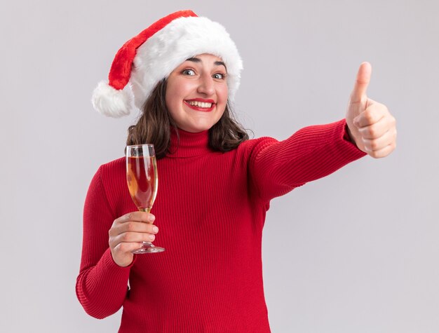 Feliz niña en suéter rojo y gorro de Papá Noel sosteniendo una copa de champán mirando a la cámara con una sonrisa en la cara mostrando los pulgares para arriba sobre fondo blanco.