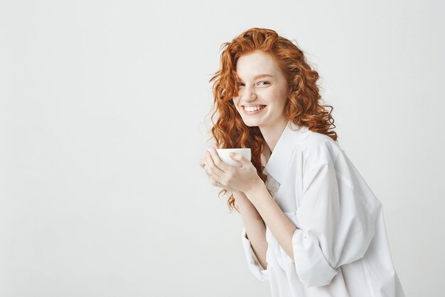 Feliz niña tierna pelirroja en camisa sonriendo sosteniendo la taza sentado en la mesa