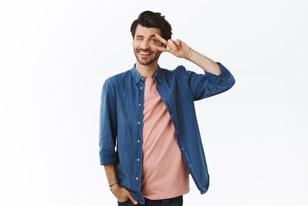 Feliz novio barbudo actuando lindo y tonto mostrando la paz o el signo de la victoria sobre un guiño de ojo descarado a la cámara y sonríe adorablemente con una mano en el bolsillo de pie con fondo blanco positivo