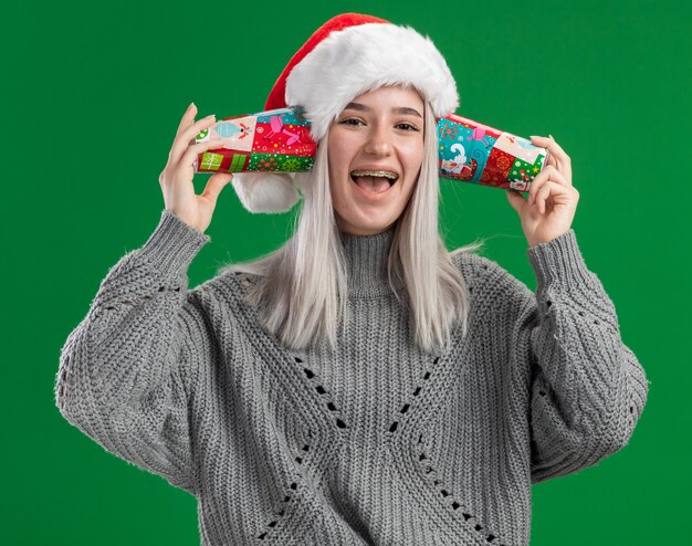Feliz oung mujer rubia en suéter de invierno y gorro de Papá Noel sosteniendo coloridos vasos de papel sobre sus orejas sonriendo alegremente de pie sobre fondo verde