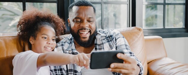 Feliz padre e hija de familia afroamericana divirtiéndose y usando videollamadas de teléfono móvil en el sofá de la casa.