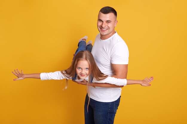 Foto gratuita feliz padre e hija de la familia, los niños desess denim onalls fingiendo ser avión con las manos extendidas a un lado y divirtiéndose con su papá, aislado en la pared amarilla