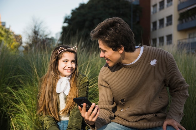 Foto gratuita feliz padre e hija mirando el uno al otro sosteniendo el teléfono móvil en la mano