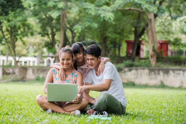 Feliz padre de familia, madre e hija sentada en la hierba y jugando la computadora portátil en el parque al aire libre