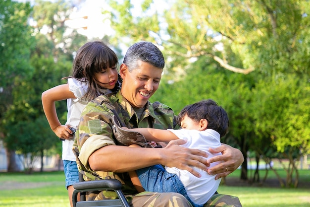 Feliz papá militar discapacitado caminando con dos niños en el parque. Niña sosteniendo asas de silla de ruedas, niño descansando sobre el regazo de papá. Veterano de guerra o concepto de discapacidad