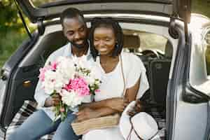 Foto gratuita feliz pareja abrazándose en el maletero del coche hatchback abierto. hombre africano abrazando a su novia. mujer sosteniendo ramo de flores, peonías.