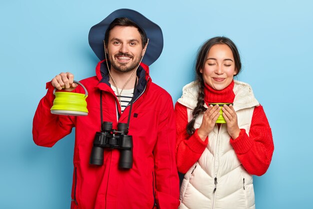 Feliz pareja de acampar bebe café temprano en la mañana, pasa tiempo libre en el campamento cerca de la hoguera, lleva binoculares, deténgase para descansar, use ropa abrigada, aislado sobre una pared azul