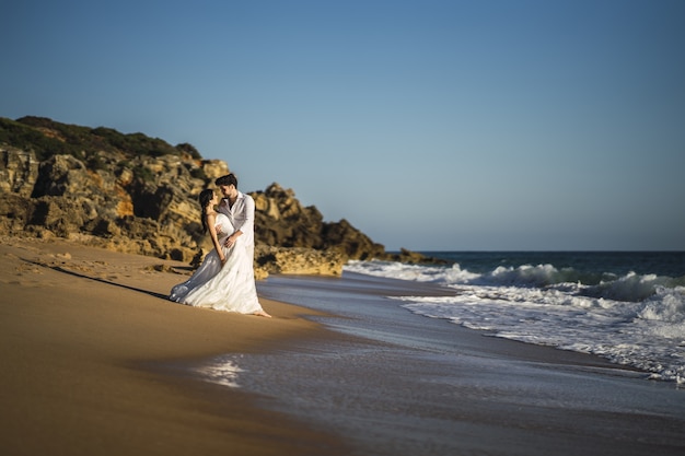 Feliz pareja amorosa caucásica vestida de blanco abrazándose en la playa durante una sesión de fotos de boda