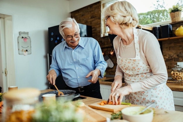 Feliz pareja de ancianos hablando y divirtiéndose mientras prepara la comida en la cocina.