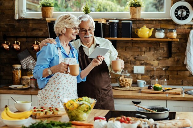 Feliz pareja de ancianos navegando por la red en el panel táctil mientras bebe café en la cocina.