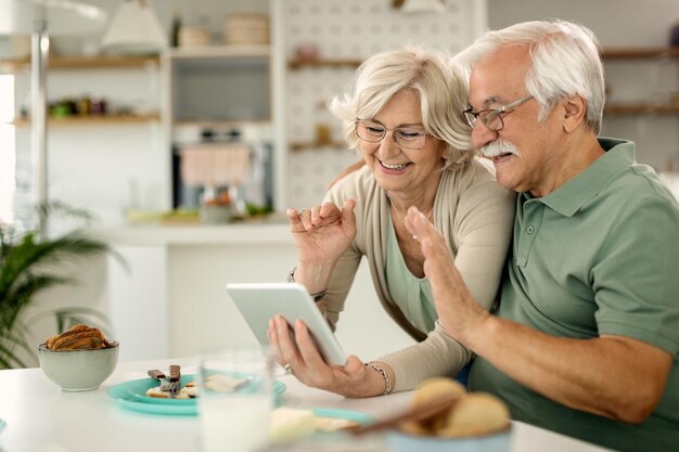Feliz pareja de ancianos saludando mientras hace una videollamada a través del panel táctil en casa