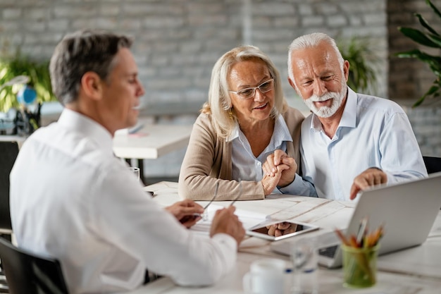 Feliz pareja de ancianos tomándose de la mano y usando una computadora portátil mientras se reúne con un asesor financiero en la oficina El hombre mayor está señalando algo en la computadora portátil