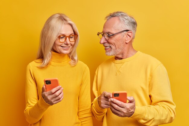 Feliz pareja de ancianos usan teléfonos móviles en casa, tienen una conversación agradable, usan ropa casual, posan contra la pared amarilla vívida