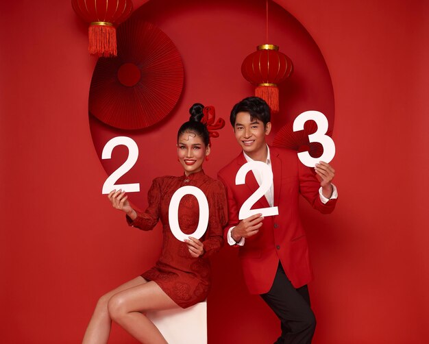 Feliz pareja asiática con atuendo informal rojo que muestra el número 2023 saludando feliz año nuevo