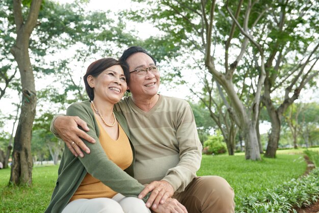 Feliz pareja asiática en fecha sentado en el banco en el parque mirando a otro lado