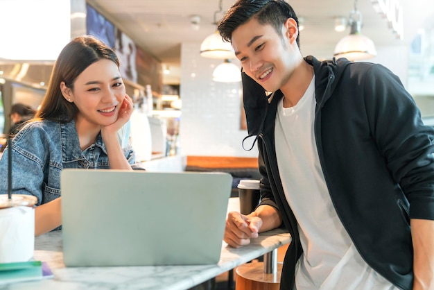 Feliz pareja asiática se quita la máscara facial en una cafetería navegando por Internet en una computadora portátil Un hombre y una mujer jóvenes en un restaurante mirando la computadora con pantalla táctil se ríen y sonríen juntos