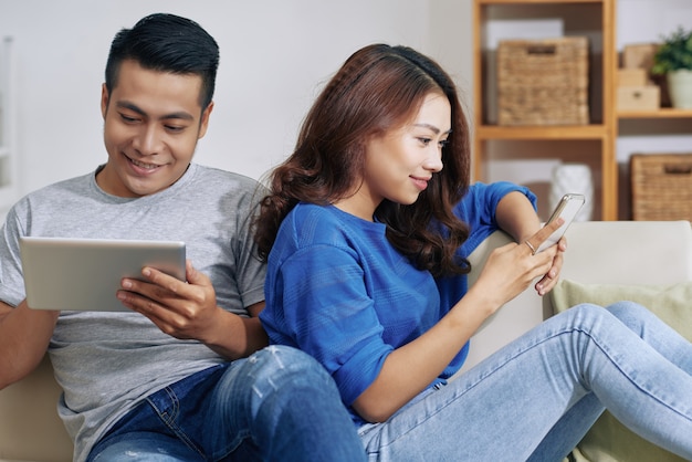 Feliz pareja asiática sentada en el sofá en casa con gadgets