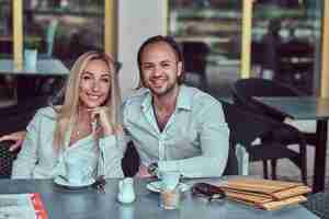 Foto gratuita feliz pareja atractiva: una encantadora mujer rubia vestida con una blusa blanca y un hombre barbudo con un elegante corte de pelo vestido con una camisa blanca durante una cita en un café al aire libre.