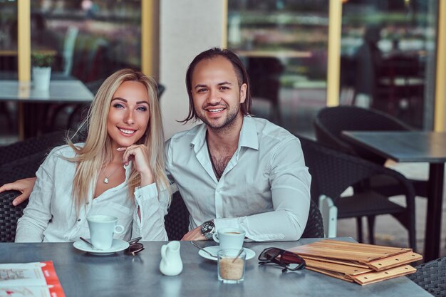 Feliz pareja atractiva: una encantadora mujer rubia vestida con una blusa blanca y un hombre barbudo con un elegante corte de pelo vestido con una camisa blanca durante una cita en un café al aire libre.