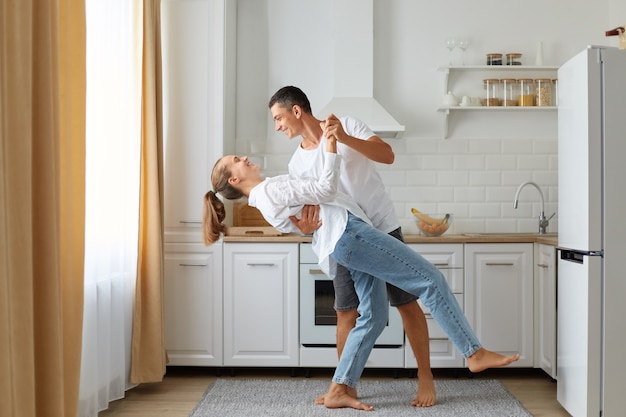 Feliz pareja bailando en la cocina, marido y mujer vestidos con camisas blancas bailan por la mañana cerca de la ventana, expresando amor y sentimientos románticos, tiro interior.