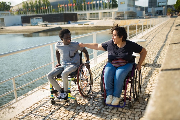 Feliz pareja birracial caminando el día de verano. Hombre afroamericano y mujer caucásica en sillas de ruedas en el terraplén, bromeando. Amor, relación, concepto de felicidad.