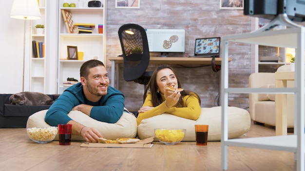 Feliz pareja caucásica comiendo pizza sentados en almohadas en el suelo viendo la televisión mientras su gato se relaja en el fondo.