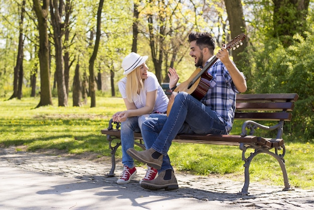 Feliz pareja caucásica sentada en un banco del parque, con el hombre tocando la guitarra durante el día
