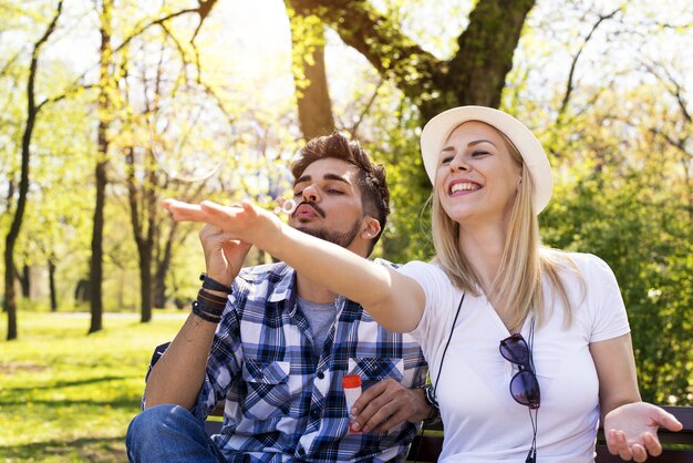 Feliz pareja caucásica sentado en un banco del parque, soplando burbujas y divirtiéndose