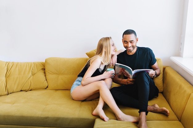 Feliz pareja descansando en casa en el sofá, pasar tiempo juntos. Empareje en ropa de casa leyendo una revista con una sonrisa en sus rostros. Comodidad. recreación, relax, comunicación.