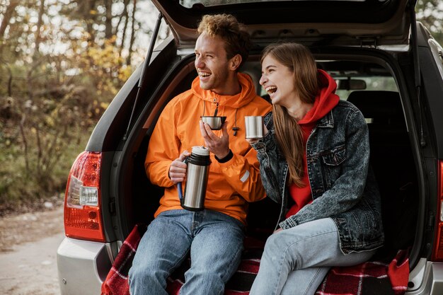 Feliz pareja disfrutando de una bebida caliente en el maletero del coche