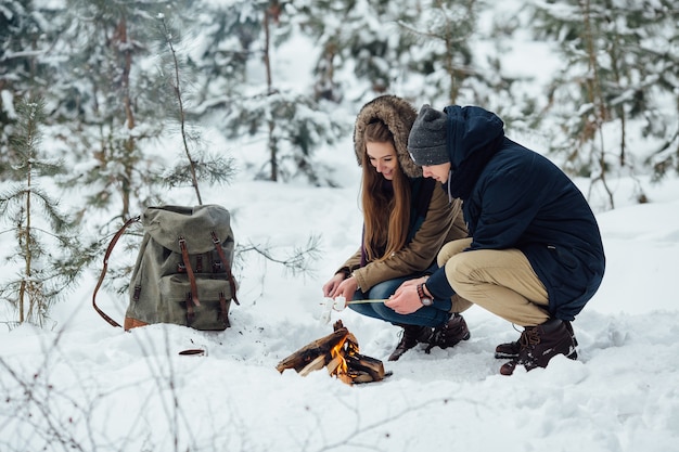 Feliz pareja de enamorados asar malvaviscos en una fogata en el clima de invierno cubierto de nieve.