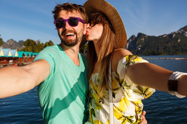 Feliz pareja encantadora haciendo selfie en vacaciones en las montañas y el lago, sombrero de ropa brillante de verano y gafas de sol, besos y diversión juntos.