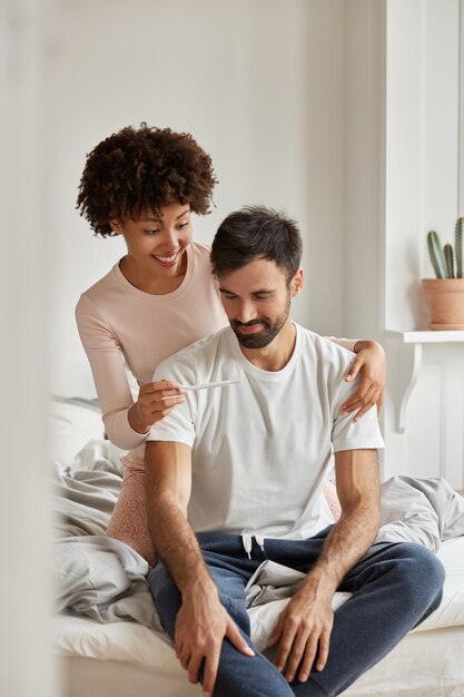 Feliz pareja de familia multiétnica mira felizmente la prueba de embarazo, se siente emocionada, celebra buenas noticias, posa en el dormitorio, usa ropa casual, se sienta en una cama cómoda durante la mañana. Concepto de fertilidad