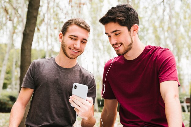 Feliz pareja gay en auriculares escuchando música en el móvil en el parque